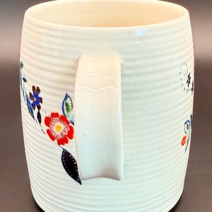 May include: A white ceramic mug with a ribbed texture and a white handle. The mug is decorated with colorful floral designs in red, blue, and green. The mug has a black base and a cream-colored interior.