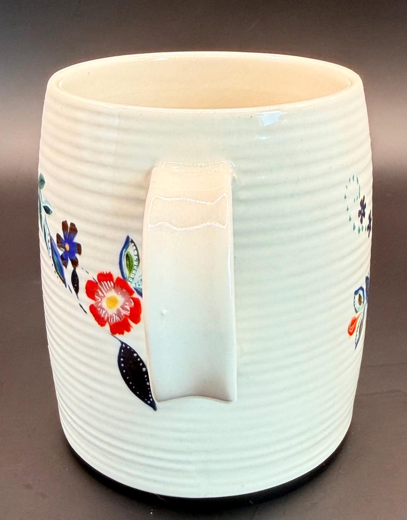 May include: A white ceramic mug with a ribbed texture and a white handle. The mug is decorated with colorful floral designs in red, blue, and green. The mug has a black base and a cream-colored interior.