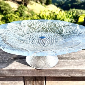 May include: A clear glass cake stand with a sunflower design. The stand has a wide, flat base and a tall, slender pedestal.