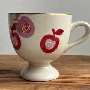 May include: A white ceramic teacup with a gold rim and a footed base. The cup is decorated with red and pink apples with a white and gold patterned design.