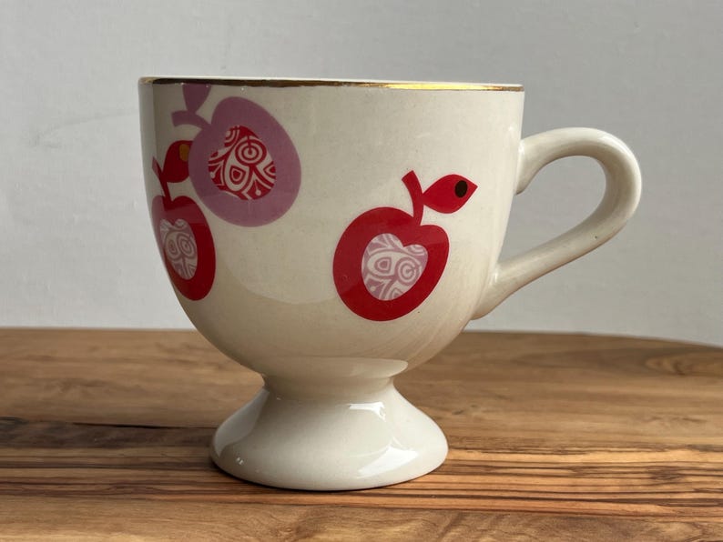 May include: A white ceramic teacup with a gold rim and a footed base. The cup is decorated with red and pink apples with a white and gold patterned design.