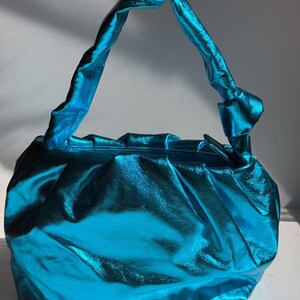 May include: A turquoise, metallic-finish handbag with a gathered design. The bag features a knotted handle and a rounded shape. The material has a shiny, reflective quality, and the bag appears to be empty.