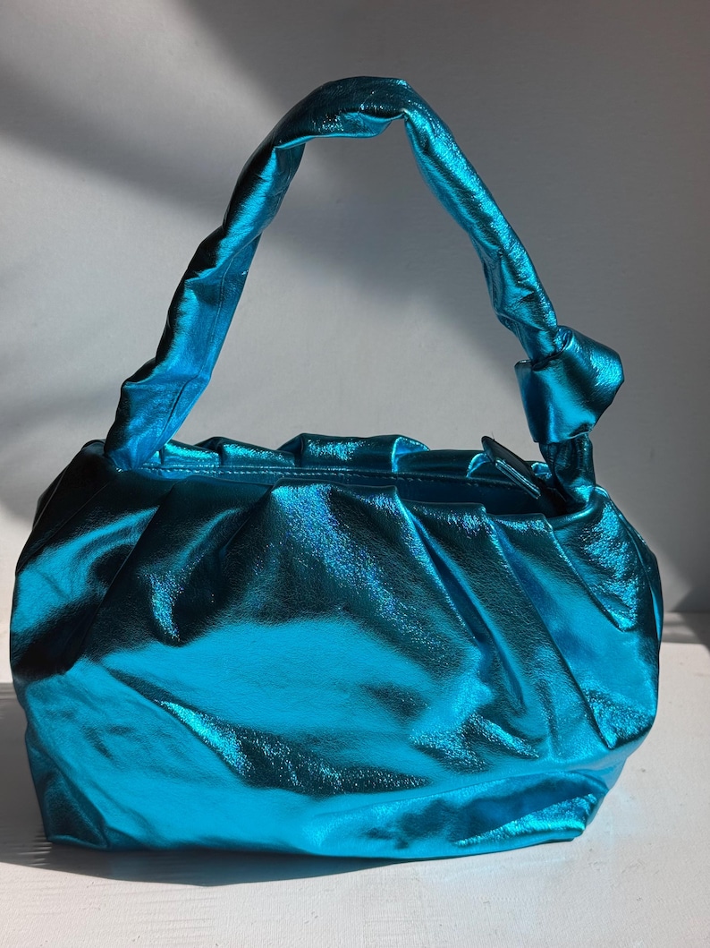 May include: A turquoise, metallic-finish handbag with a gathered design. The bag features a knotted handle and a rounded shape. The material has a shiny, reflective quality, and the bag appears to be empty.