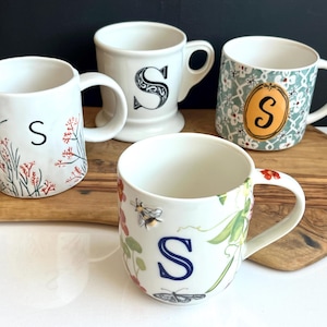 Pu&ograve; includere: Quattro tazze in ceramica bianca con la lettera "S". Una presenta decorazioni floreali e rosse, un'altra una "S" nera, una terza un motivo floreale con un ovale arancione e l'ultima un design con api e farfalle.