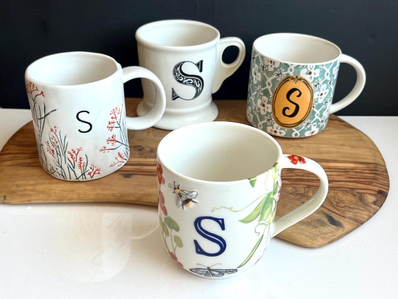 Pu&ograve; includere: Quattro tazze in ceramica bianca con la lettera "S". Una presenta decorazioni floreali e rosse, un'altra una "S" nera, una terza un motivo floreale con un ovale arancione e l'ultima un design con api e farfalle.