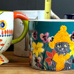 May include: A selection of ceramic mugs with various designs. One mug has a parrot design with green, white, orange, and red details. Another features a cat wearing a yellow hat and scarf, surrounded by flowers. A blue mug with a gold handle is also present.