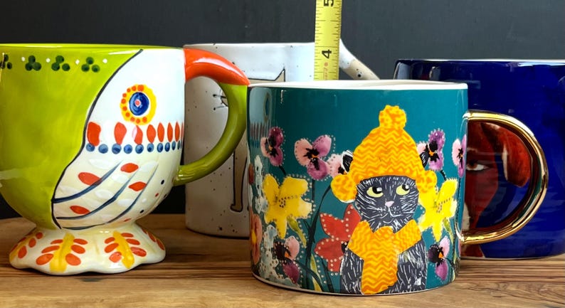 May include: A selection of ceramic mugs with various designs. One mug has a parrot design with green, white, orange, and red details. Another features a cat wearing a yellow hat and scarf, surrounded by flowers. A blue mug with a gold handle is also present.