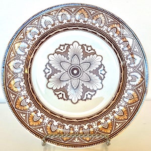 May include: A white ceramic plate with a brown and gold floral design. The plate has a raised rim and a center design with a brown and white floral pattern.