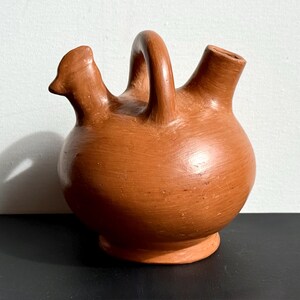 May include: A terracotta-colored ceramic pitcher with a rounded body, a handle, and two spouts. One spout is shaped like a bird's head. The pitcher sits on a small, circular base.