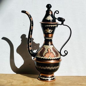 May include: A black and copper teapot with intricate floral designs. The teapot has a long, curved spout and a handle with a decorative curl.