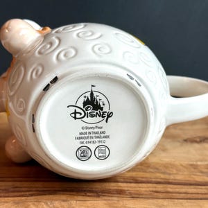 May include: White ceramic Disney mug with a sheep design, featuring embossed details and a curved handle. The mug's base displays the Disney logo and text. The mug is resting on a wooden surface.