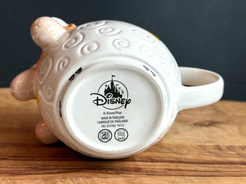 May include: White ceramic Disney mug with a sheep design, featuring embossed details and a curved handle. The mug's base displays the Disney logo and text. The mug is resting on a wooden surface.