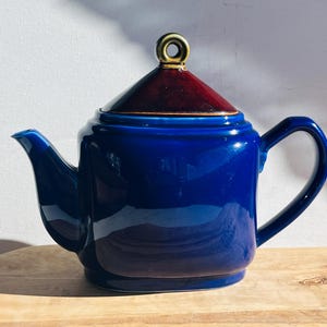 May include: A blue ceramic teapot with a burgundy lid and gold accents. The teapot has a square body and a curved spout.