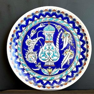 May include: A round, ceramic decorative plate with a white rim and a vibrant blue center. The plate features a detailed design of a vase and floral motifs in shades of blue, turquoise, and white. The outer border has a repeating pattern.