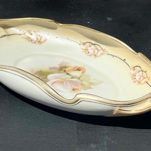 May include: Oval-shaped, antique porcelain dish with a cream-colored interior and a pale yellow exterior. The dish features a floral rose design in the center and along the rim, with gold trim. It has handles on each end.