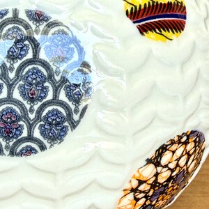 May include: Close-up of a white ceramic plate with a wavy texture. The plate features three decorative, colorful, oval-shaped designs. One design has a blue and purple floral pattern, and the other two have vibrant, patterned fabric designs.