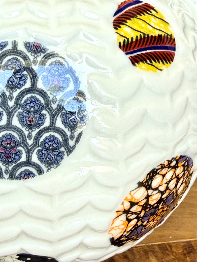 May include: Close-up of a white ceramic plate with a wavy texture. The plate features three decorative, colorful, oval-shaped designs. One design has a blue and purple floral pattern, and the other two have vibrant, patterned fabric designs.