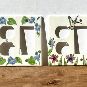May include: Two white ceramic light switch covers with floral designs. One cover features a hummingbird and the other has blue flowers.