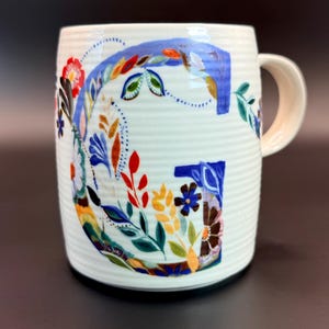May include: White ceramic mug with a large, colorful floral "G" design. The letter is filled with blue, red, yellow, and green flowers and leaves. The mug has a ribbed texture and a cream-colored handle.