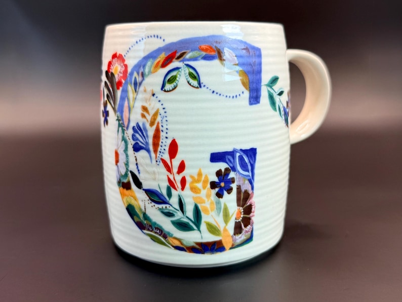 May include: White ceramic mug with a large, colorful floral "G" design. The letter is filled with blue, red, yellow, and green flowers and leaves. The mug has a ribbed texture and a cream-colored handle.