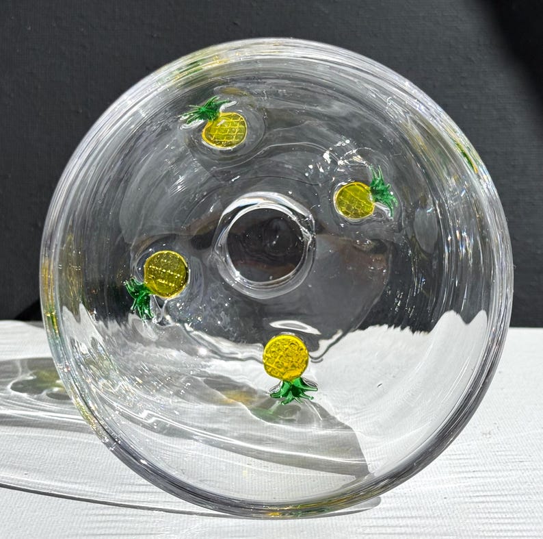 May include: Clear glass bowl with a central hole and decorative yellow and green pineapple designs. The bowl is round and has a slightly curved shape, with the pineapple designs evenly spaced around the interior.