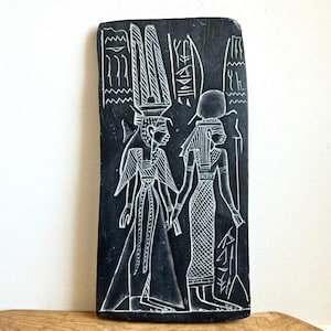 May include: A black rectangular stone carving featuring two figures in ancient Egyptian attire, holding hands. The figures are depicted in white relief, with hieroglyphs and ornate headdresses. The carving is set against a neutral background.