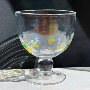 May include: Clear glass goblet with a wide bowl and a short stem. The bowl is decorated with yellow and green pineapple designs. The goblet sits on a round base and has a small spout on the rim.