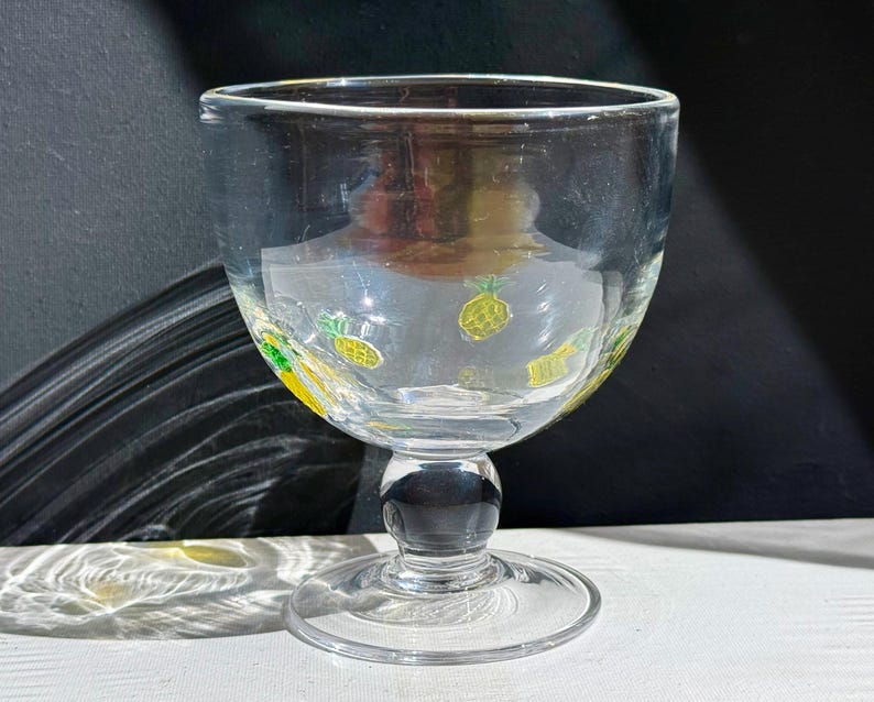 May include: Clear glass goblet with a wide bowl and a short stem. The bowl is decorated with yellow and green pineapple designs. The goblet sits on a round base and has a small spout on the rim.