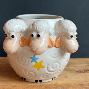 May include: A white ceramic sheep-themed container with three sheep heads protruding from the rim. The container features a blue star and a yellow star design, with black feet. The sheep have pink faces and orange accents.
