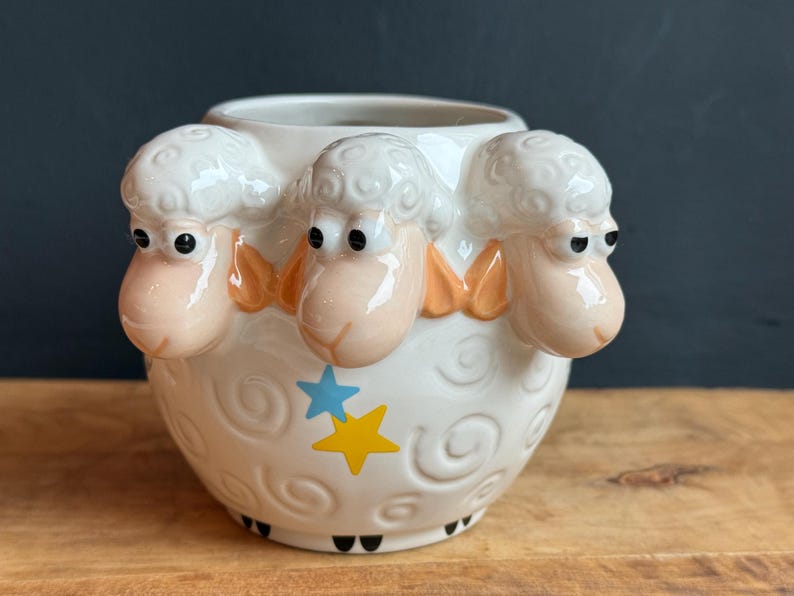 May include: A white ceramic sheep-themed container with three sheep heads protruding from the rim. The container features a blue star and a yellow star design, with black feet. The sheep have pink faces and orange accents.