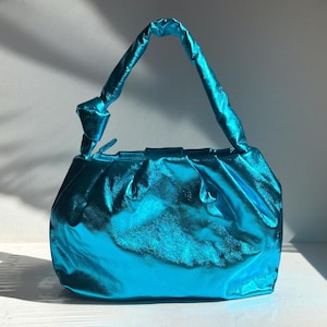 May include: A turquoise metallic handbag with a knotted handle. The bag has a gathered design and a shiny, reflective surface. The handle is a twisted, tube-like shape. The bag is sitting on a white surface.