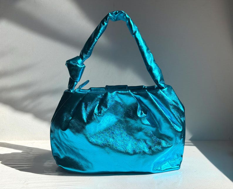 May include: A turquoise metallic handbag with a knotted handle. The bag has a gathered design and a shiny, reflective surface. The handle is a twisted, tube-like shape. The bag is sitting on a white surface.