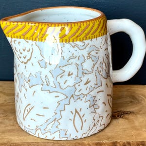 May include: A ceramic pitcher with a white base, adorned with a pattern of light blue and brown leaf-like designs. The rim features a yellow band with brown diagonal stripes. It has a white, rounded handle.