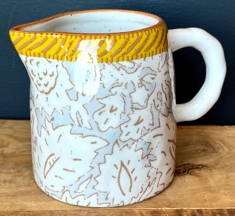 May include: A ceramic pitcher with a white base, adorned with a pattern of light blue and brown leaf-like designs. The rim features a yellow band with brown diagonal stripes. It has a white, rounded handle.