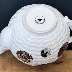 May include: White ceramic teapot with a textured, patterned surface. The teapot features a curved spout and handle, and decorative oval patches in shades of brown, black, and purple. The bottom of the teapot has a circular logo with a bird and the text "MADE IN CHINA FABRIQUE EN CHINE".