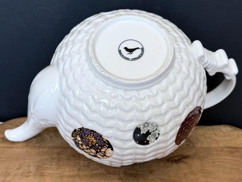 May include: White ceramic teapot with a textured, patterned surface. The teapot features a curved spout and handle, and decorative oval patches in shades of brown, black, and purple. The bottom of the teapot has a circular logo with a bird and the text "MADE IN CHINA FABRIQUE EN CHINE".
