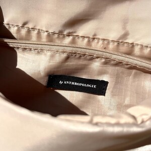 K&ouml;nnte beinhalten: Nahaufnahme des Inneren einer beigefarbenen Tasche mit Rei&szlig;verschluss und einem schwarzen Etikett mit der Aufschrift "by ANTHROPOLOGIE". Der Stoff hat einen leichten Glanz. Die Beleuchtung hebt die Textur und Farbe des Materials hervor.