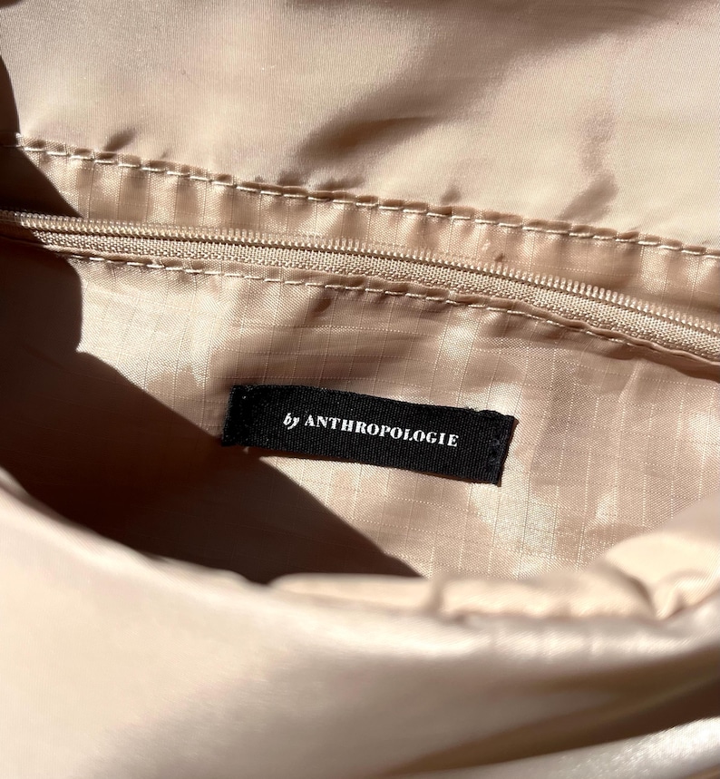 K&ouml;nnte beinhalten: Nahaufnahme des Inneren einer beigefarbenen Tasche mit Rei&szlig;verschluss und einem schwarzen Etikett mit der Aufschrift "by ANTHROPOLOGIE". Der Stoff hat einen leichten Glanz. Die Beleuchtung hebt die Textur und Farbe des Materials hervor.
