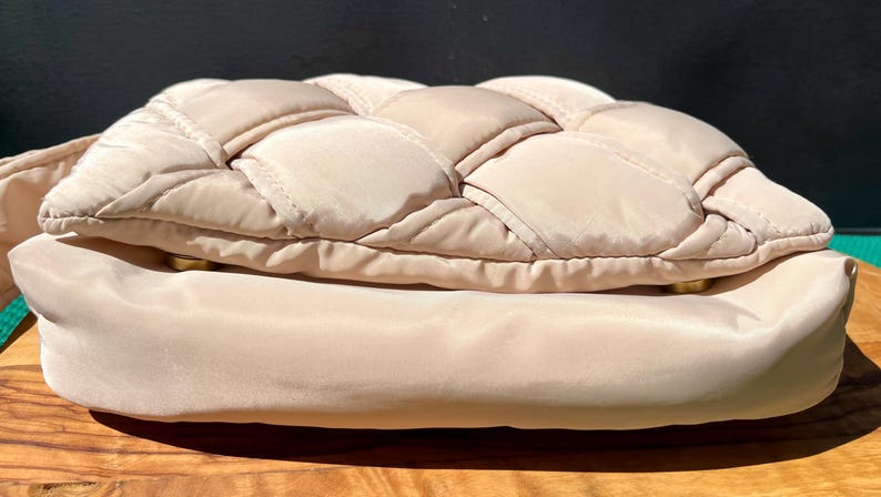 K&ouml;nnte beinhalten: Eine beige gesteppte Handtasche mit einem gewebten Design auf der oberen Klappe. Die Tasche hat eine glatte, abgerundete Basis und goldfarbene Hardware. Die Tasche ist aus einem weichen, gepolsterten Material gefertigt.