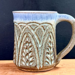 May include: A handmade ceramic mug with a light blue rim and handle. The body of the mug features a detailed, carved design in shades of green and brown. The mug is designed for hot beverages.