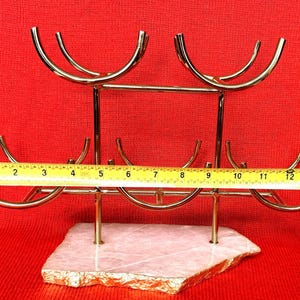 May include: A gold-coloured metal wine rack with a natural stone base. The rack holds six bottles and measures approximately 33 cm wide. The base is a light pink colour with gold edging, set against a red background.