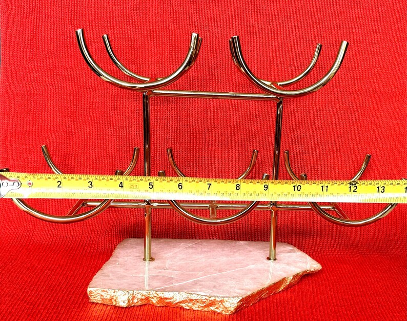 May include: A gold-coloured metal wine rack with a natural stone base. The rack holds six bottles and measures approximately 33 cm wide. The base is a light pink colour with gold edging, set against a red background.