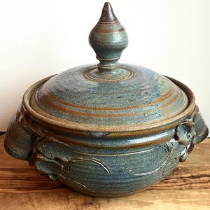 May include: A blue and brown ceramic casserole dish with a lid and decorative handles. The dish has a glazed finish and a rustic look.