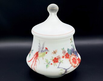 May include: White frosted glass jar with a lid, featuring a hand-painted design of a bird, flowers, and butterflies in red, green, and blue. The jar has a rounded shape and a clear glass knob on top.