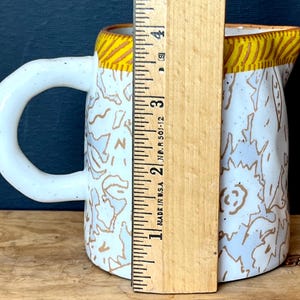 May include: White ceramic pitcher with a large handle, decorated with brown and white abstract patterns. The rim has a yellow and brown striped design. The pitcher is approximately 4 inches tall.