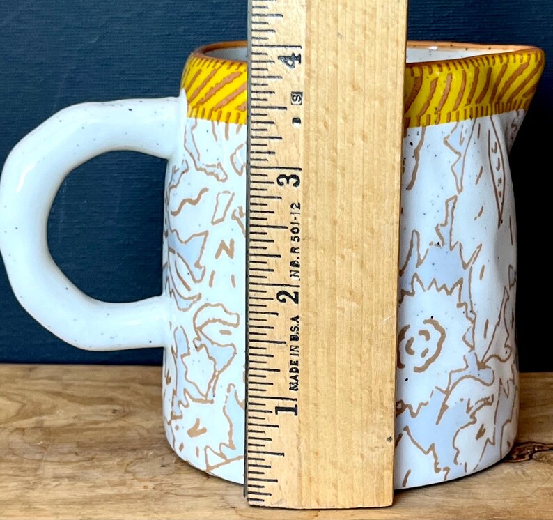 May include: White ceramic pitcher with a large handle, decorated with brown and white abstract patterns. The rim has a yellow and brown striped design. The pitcher is approximately 4 inches tall.