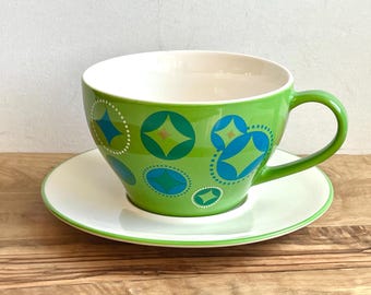 May include: A large green teacup and saucer set. The cup features a repeating pattern of blue and green geometric shapes with white dotted outlines. The saucer is white with a green trim. The cup has a large handle.