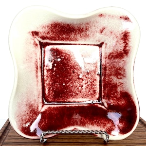 May include: A white ceramic square dish with a red glaze. The dish has a slightly raised rim and a scalloped edge. The glaze is uneven, creating a mottled effect.