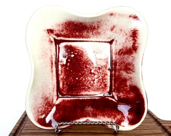 May include: A white ceramic square dish with a red glaze. The dish has a slightly raised rim and a scalloped edge. The glaze is uneven, creating a mottled effect.