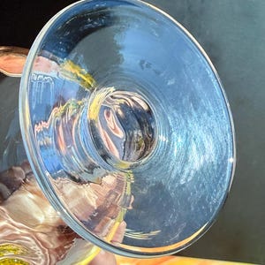May include: A clear glass goblet with a wide, shallow bowl. The glass reflects light, creating a blue hue. The stem is not visible. The goblet is held by a person.
